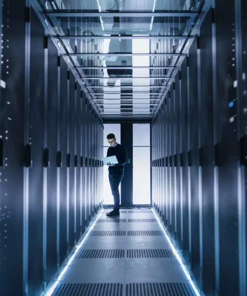 Male IT Engineer Works on a Laptop in a Big Data Center. Rows of Rack Servers are Seen.